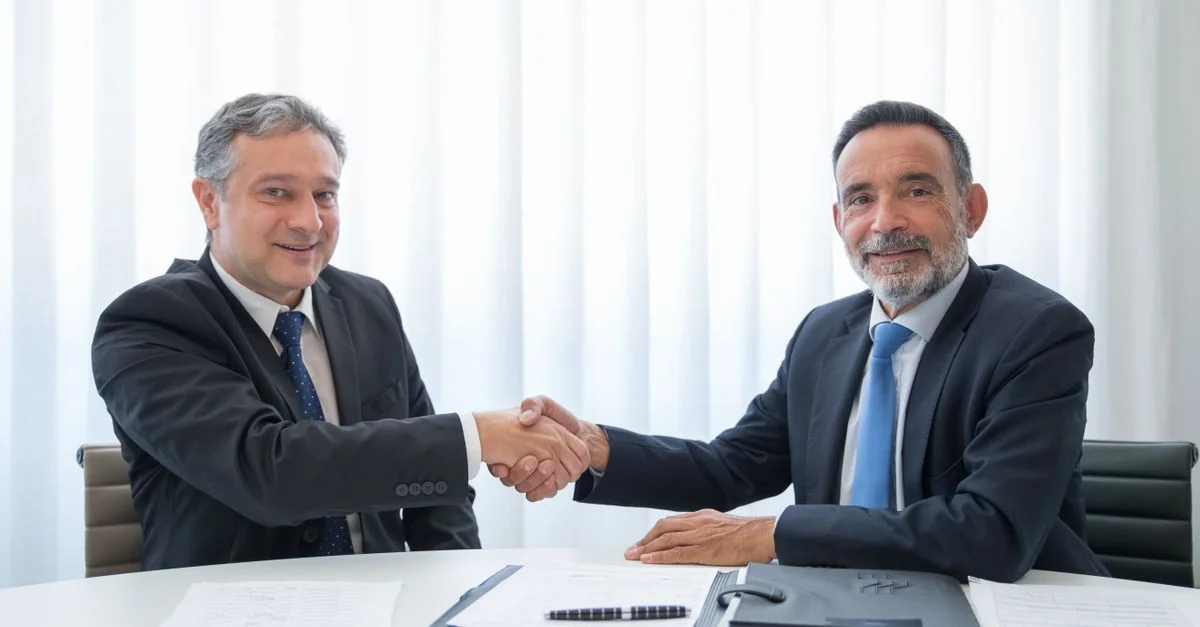 Deux hommes d'affaires en costumes se serrant la main à une table de bureau avec des documents.