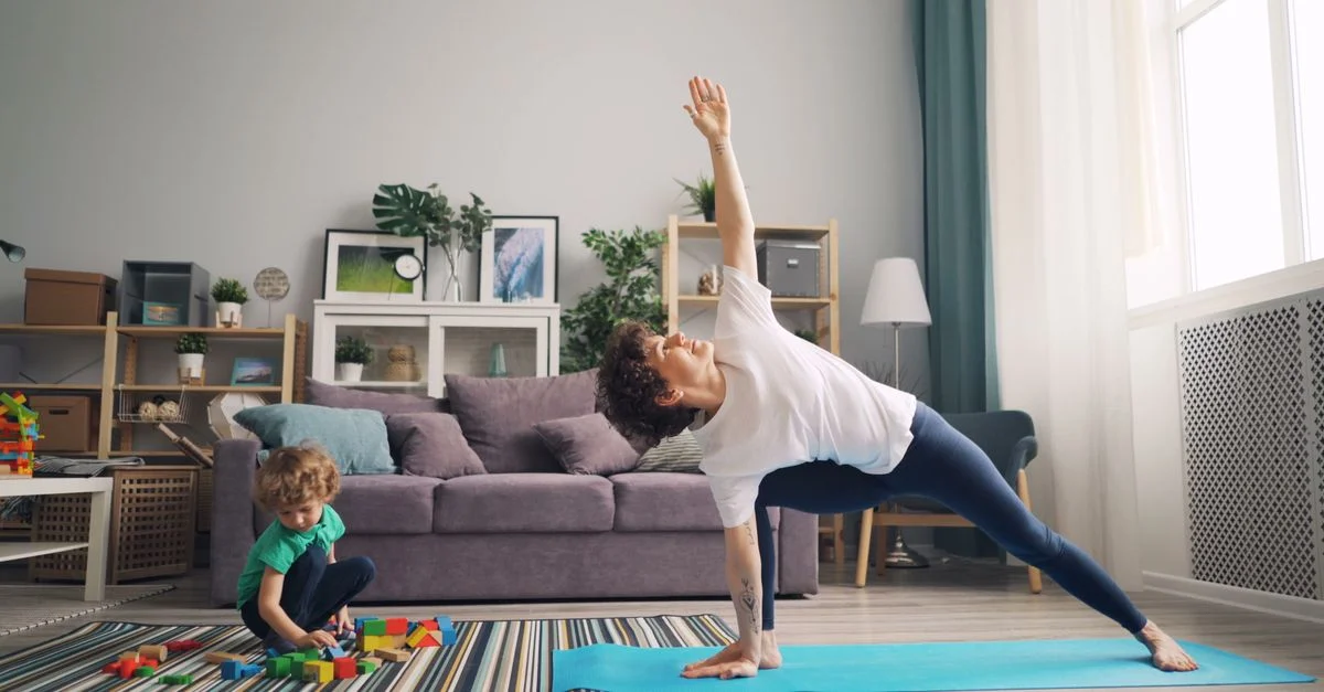 Une mère s'étire sur un tapis de yoga pendant que son enfant joue avec des blocs à proximité dans leur salon.